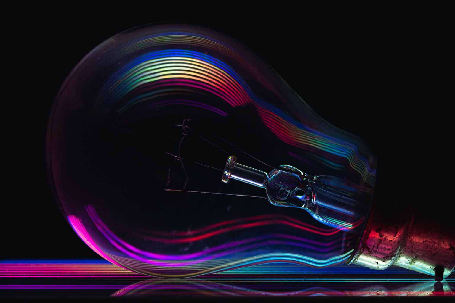 An offline, unlit lightbulb set against a deep, dark background whose clear glass catches a spectrum of rainbow hues