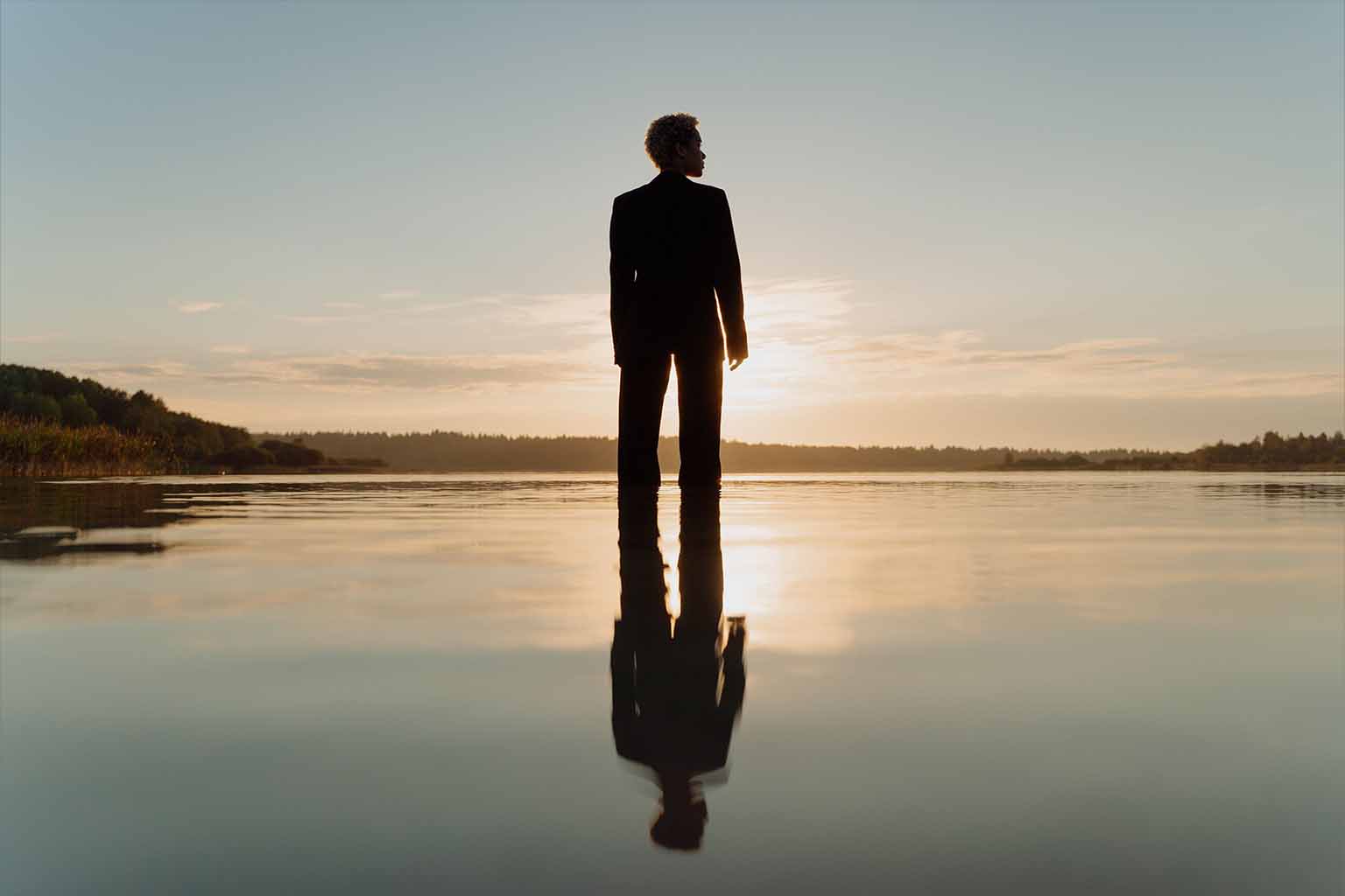 A peaceful image of person in a suit standing in shallow water in front of a sunset.